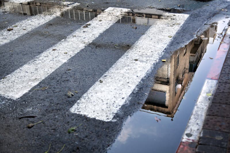 A Tel-Aviv building reflecting in a puddle that's between the curb and the crosswalk zebra stripes. Winter. A Tel-Aviv building reflecting in a puddle that's between the curb and the crosswalk zebra stripes. Winter.