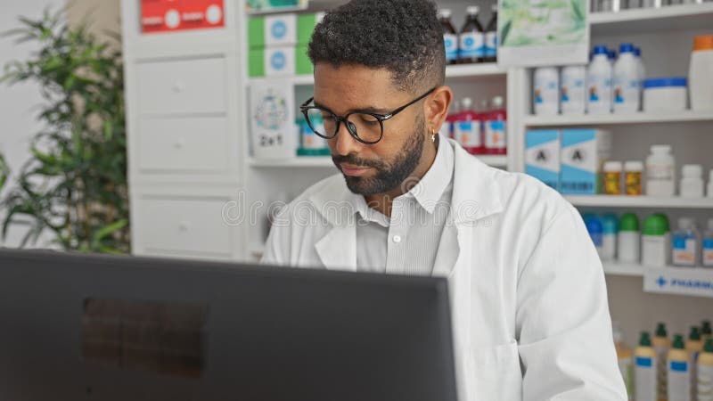 Un bărbat afro-american profesionist concentrat într-o haină albă lucrează la un computer într-o farmacie bine aprovizionată stock photos