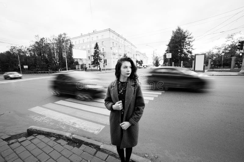 Un cappotto blu dell'adolescente che sta al semaforo sulla via della città cloudly un giorno di autunno con i veicoli che passano fotografie stock Un cappotto blu dell'adolescente che sta al semaforo sulla via della città cloudly un giorno di autunno con i veicoli che passano fotografie stock
