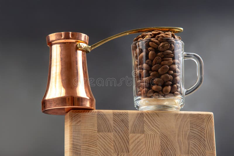 Un cezve tradițional din cupru și o cană de sticlă umplută cu boabe de cafea prăjită stau pe o suprafață din lemn pe un fond albas royalty free stock photo