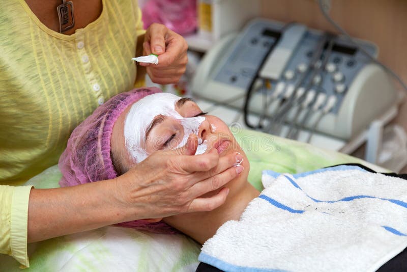 Un cosmetologist experimentado aplica una m?scara de la crema blanca en la cara de una chica joven que mienta en el sof? durante imagenes de archivo Un cosmetologist experimentado aplica una m?scara de la crema blanca en la cara de una chica joven que mienta en el sof? durante imagenes de archivo