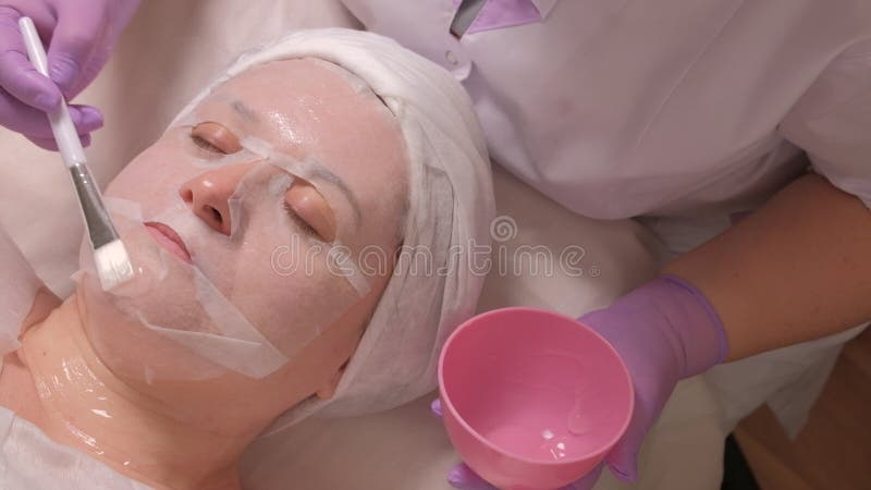 Un cosmetologist profesional en guantes de la lila mancha la crema de alimentación en la cara femenina de mediana edad Una mujer imágenes de archivo libres de regalías Un cosmetologist profesional en guantes de la lila mancha la crema de alimentación en la cara femenina de mediana edad Una mujer imágenes de archivo libres de regalías