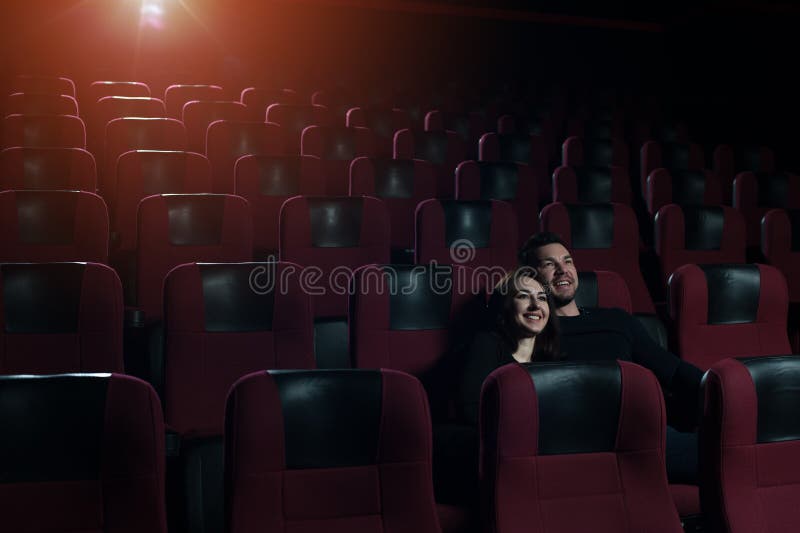 A couple in love sitting in empty movie theater. Young handsome adults watching film, men hugging his brunette girlfriend. Cinema, entertainment and leisure concept. A couple in love sitting in empty movie theater. Young handsome adults watching film, men hugging his brunette girlfriend. Cinema, entertainment and leisure concept.