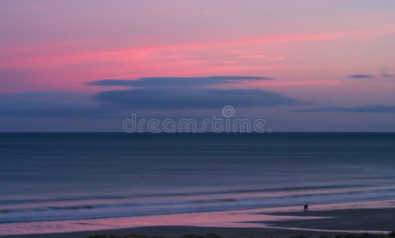 A romantic couple walk an empty beach as the gentle tide washes in against a beautiful pink and purple sunset in the last hour of light. Bamburgh, Northumberland, England, UK. A romantic couple walk an empty beach as the gentle tide washes in against a beautiful pink and purple sunset in the last hour of light. Bamburgh, Northumberland, England, UK