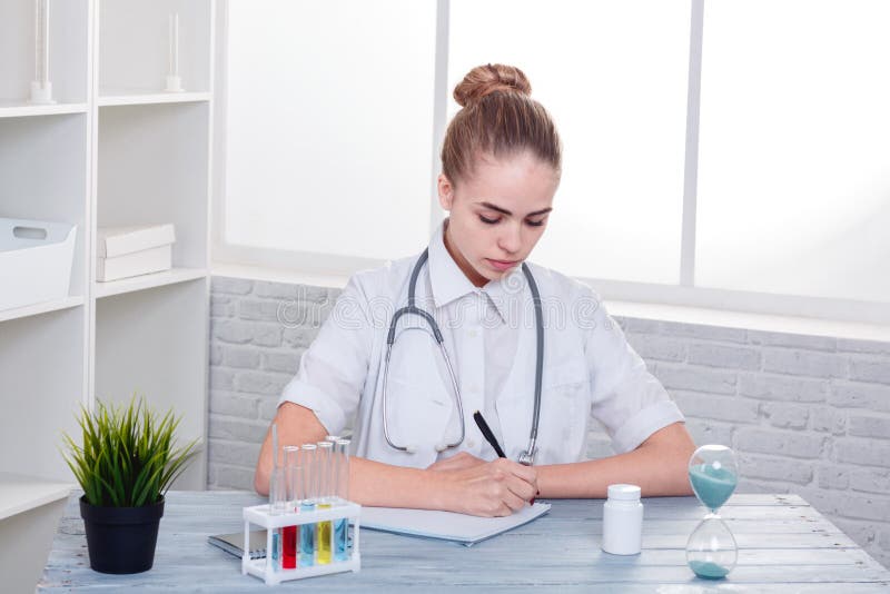 Un docteur de fille, dans l'uniforme blanc, s'assied à son bureau et écrit quelque chose à son carnet photo libre de droits Un docteur de fille, dans l'uniforme blanc, s'assied à son bureau et écrit quelque chose à son carnet photo libre de droits
