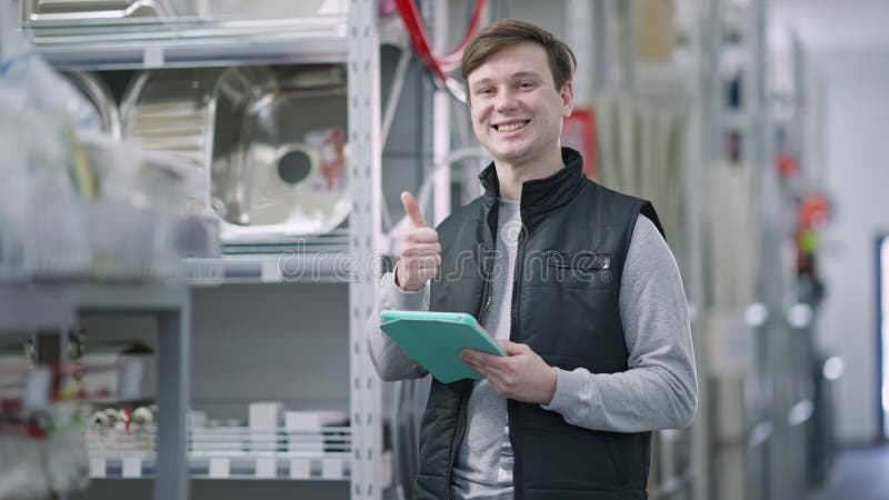 Un employé souriant et positif regarde la caméra montrant le pouce levé comme client passant à l'arrière-plan dans la quincailleri clips vidéos