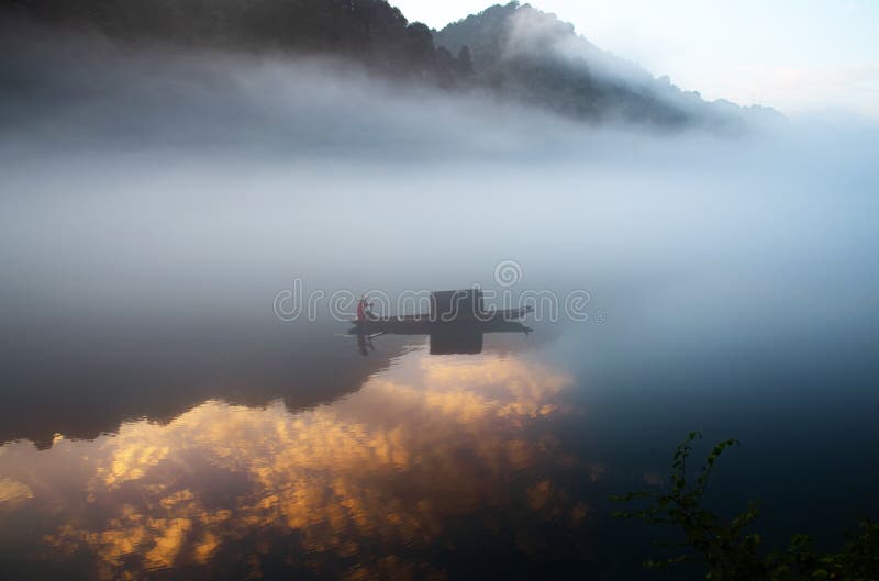 Un fishman sulla barca nella nebbia sul fiume, la riflessione dorata della nuvola sulla superficie di acqua, all'alba immagini stock Un fishman sulla barca nella nebbia sul fiume, la riflessione dorata della nuvola sulla superficie di acqua, all'alba immagini stock
