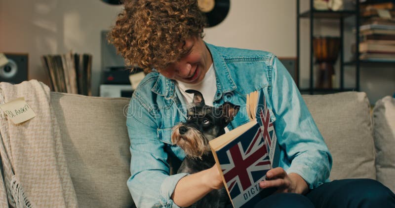 Un garçon avec un chien sur les genoux lit un dictionnaire anglais étudiant pour un test embrassant le chien fourré qui est banque de vidéos