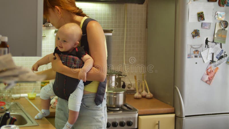 Un garçon d'enfant assis dans un support de bébé en regardant autour de moi et souriant, tandis que sa jeune mère brune à faire de