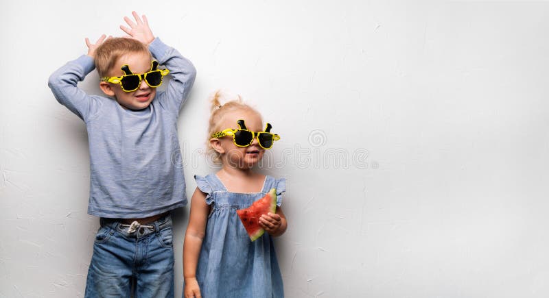 Un garçon et une fille heureux dans des lunettes de soleil jaunes mangent une pastèque rouge mûre sur une fan blanche de mur Port images stock Un garçon et une fille heureux dans des lunettes de soleil jaunes mangent une pastèque rouge mûre sur une fan blanche de mur Port images stock