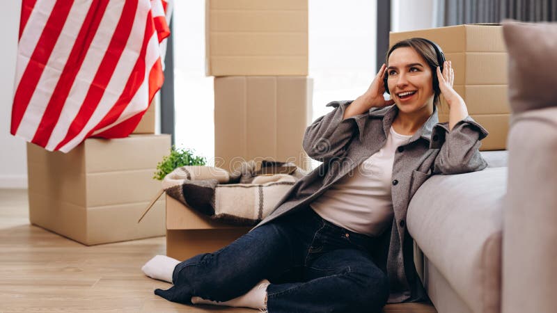 A young teenager has moved to a new apartment and is listening to music in headphones. The concept of happy moving, studentship. The USA flag is in the background. A young teenager has moved to a new apartment and is listening to music in headphones. The concept of happy moving, studentship. The USA flag is in the background.