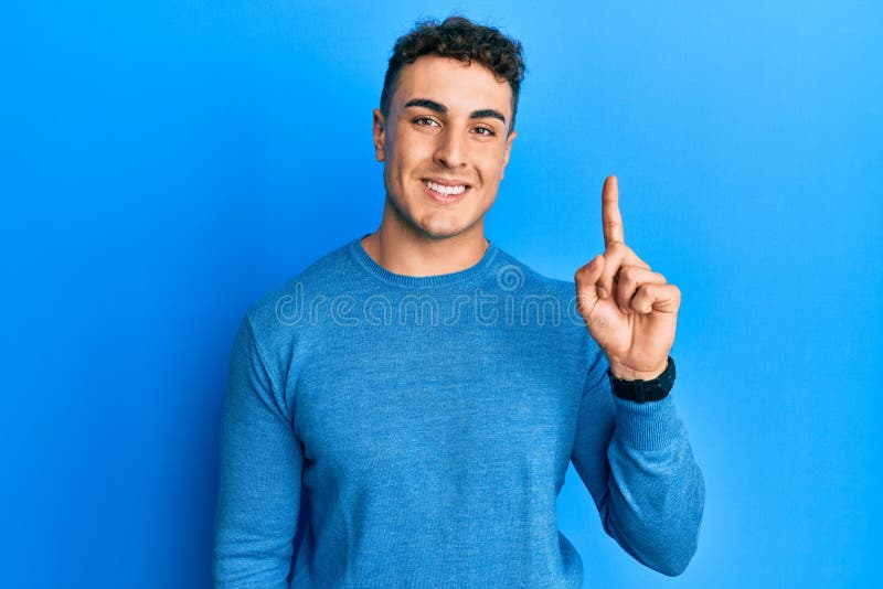 Un giovane ispanico con un maglione invernale casuale che mostra e punta il dito contro il dito numero uno, sorridendo sicuro e fe fotografie stock libere da diritti