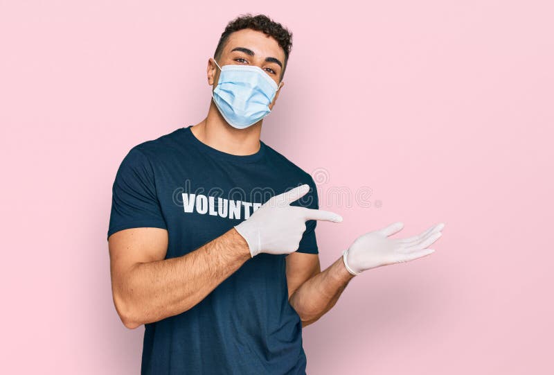 Un giovane ispanico, con una maschera medica e una camicia volontaria, sorpreso e sorridente alla telecamera mentre si presenta a fotografia stock