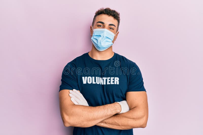 Un giovane ispanico, con una maschera medica e una maglietta volontaria, ha un viso felice sorridendo con le braccia incrociate ch fotografie stock