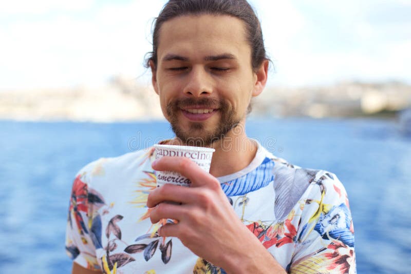 Un giovane uomo bello che mangia caffè caldo fuori vicino al mare che gode di questo momento fotografia stock Un giovane uomo bello che mangia caffè caldo fuori vicino al mare che gode di questo momento fotografia stock