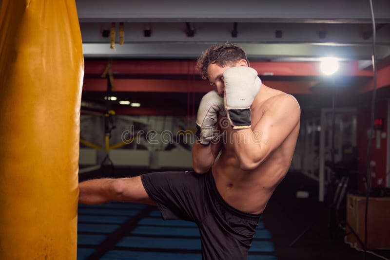 Un giovane uomo senza camicia, gamba del pugile che colpisce punching ball, practic immagini stock libere da diritti Un giovane uomo senza camicia, gamba del pugile che colpisce punching ball, practic immagini stock libere da diritti