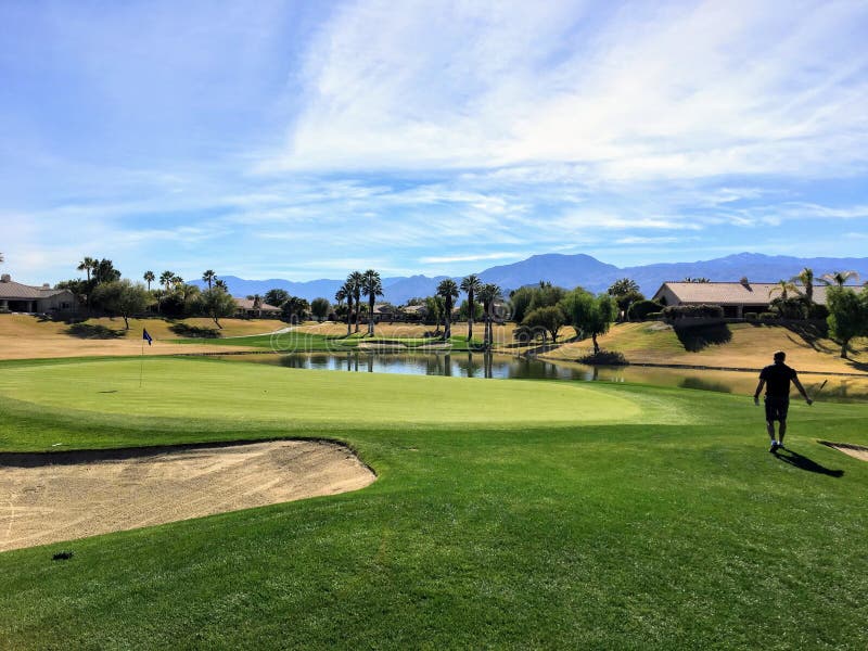 Un golfista de sexo masculino joven que camina hacia el verde en un par 4 rodeado por el agua y las palmeras en el fondo en el oa imagenes de archivo Un golfista de sexo masculino joven que camina hacia el verde en un par 4 rodeado por el agua y las palmeras en el fondo en el oa imagenes de archivo