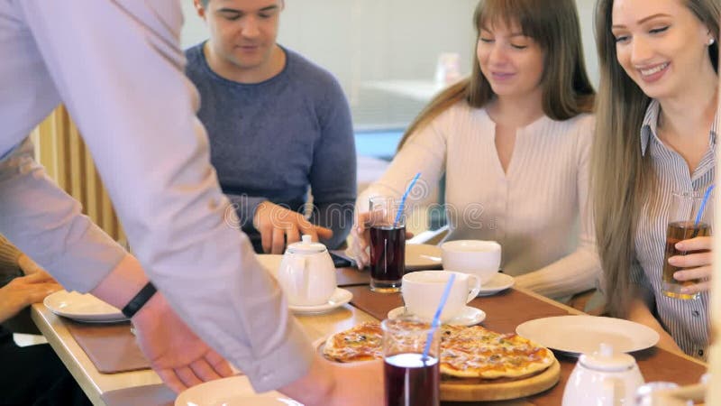 Un groupe d'amis heureux s'installe à table dans un restaurant cosy riant et s'amusant. le serveur sert de la pizza banque de vidéos