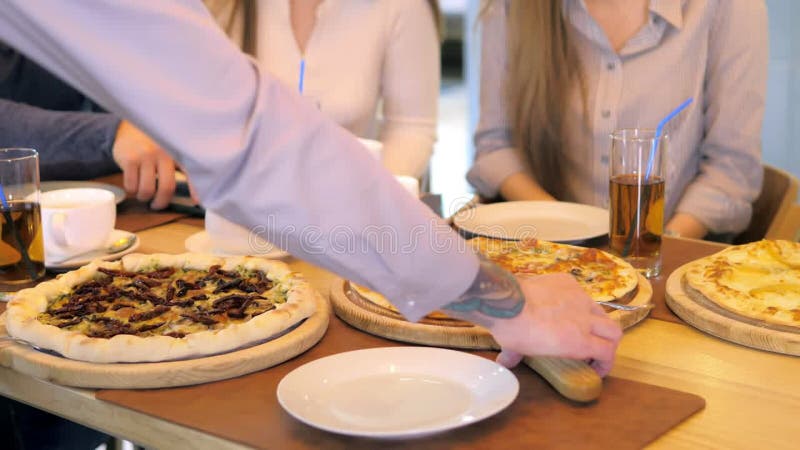 Un groupe d'amis heureux s'installe à table dans un restaurant cosy riant et s'amusant. le serveur sert de la pizza clips vidéos