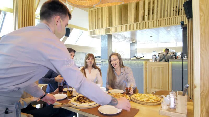 Un groupe d'amis heureux s'installe à table dans un restaurant cosy riant et s'amusant. le serveur sert de la pizza clips vidéos