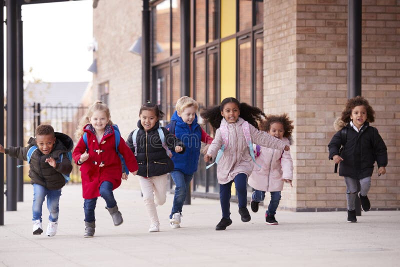 Un gruppo multi-etnico felice di giovani bambini della scuola che portano i cappotti e che portano le cartelle che si dirige in u fotografia stock Un gruppo multi-etnico felice di giovani bambini della scuola che portano i cappotti e che portano le cartelle che si dirige in u fotografia stock
