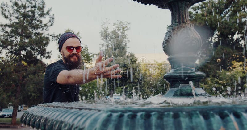 Un hombre barbudo con gafas de sol admira una fuente en un parque almacen de metraje de vídeo
