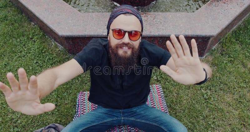 Un hombre barbudo con gafas de sol sentado sobre la hierba junto a una fuente disfrutando de la paz metrajes