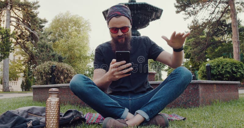 Un hombre barbudo está sentado en el pasto mirando su teléfono almacen de video