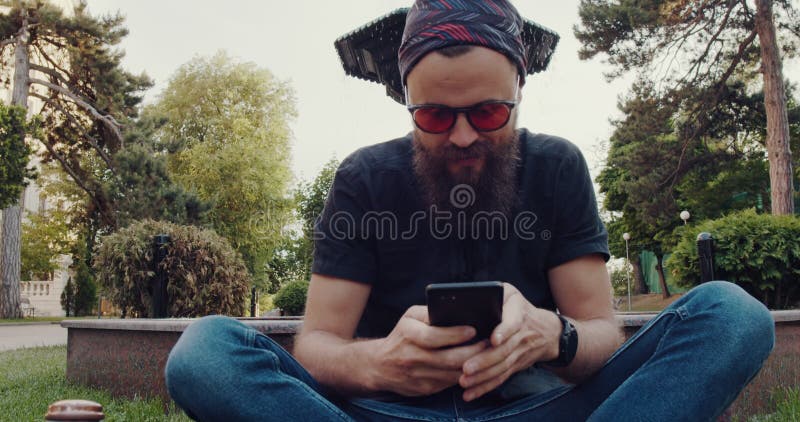 Un hombre barbudo se sienta en la hierba centrada en su celular almacen de video