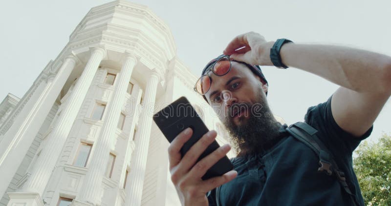 Un hombre barbudo usa su teléfono de un edificio alto que combina tecnología con vida en la ciudad almacen de metraje de vídeo
