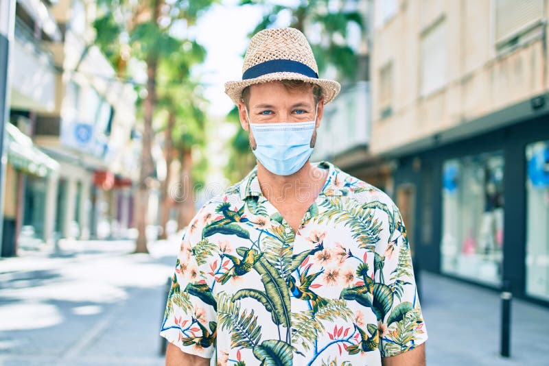 Un hombre caucásico guapo que lleva sombrero de verano y camisa floral con máscara de seguridad coronavirus al aire libre foto de archivo libre de regalías