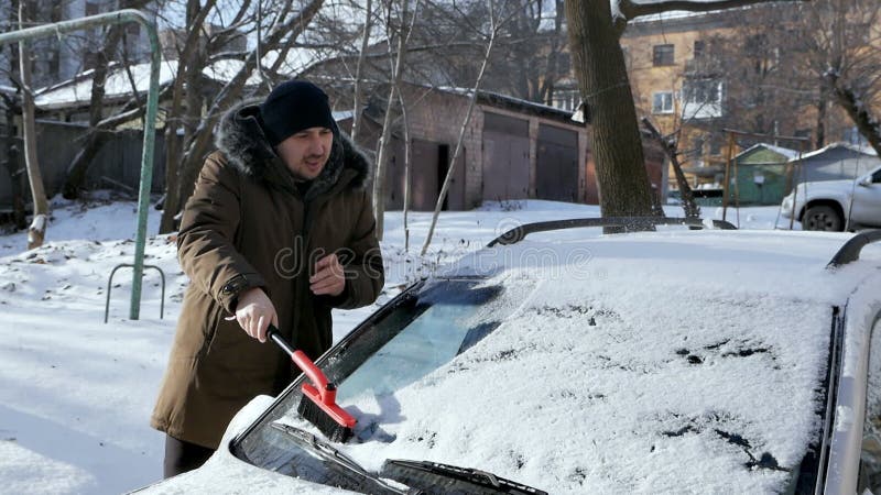 Un hombre cepilla de nieve el parabrisas de un coche. en invierno el coche estaba cubierto de nieve metrajes