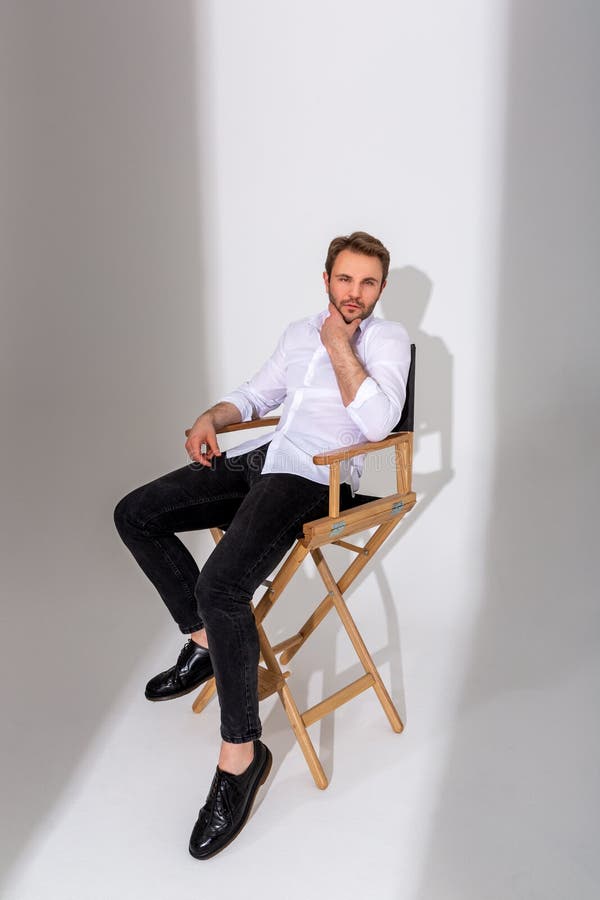 Un Hombre Con Camisa Blanca Jeans Oscuros Y Botas Negras Sentado En Una Silla Alta Contra Un Común. Desgaste Casual. Mirada Foto de archivo - Imagen de atractivo, negro: