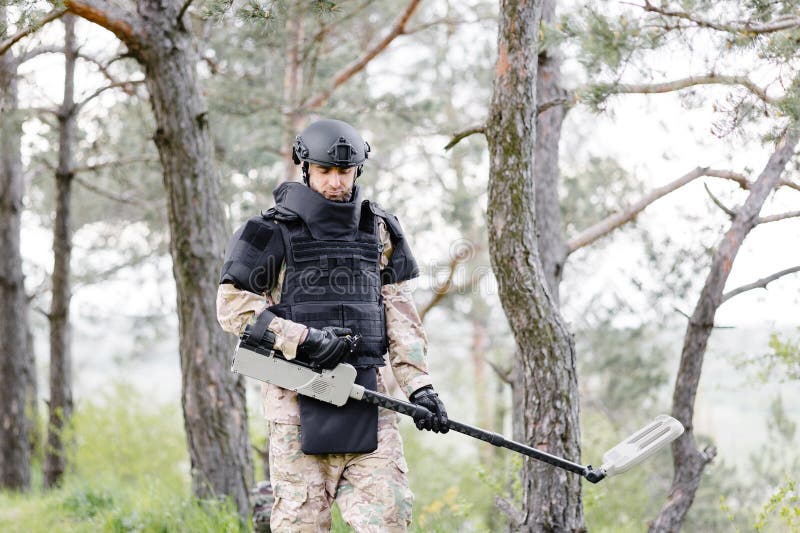 Un hombre con un uniforme militar y un chaleco antibalas se sienta en el  bosque cerca de un detector de metales y una mochila militar. Un hombre se  detiene en el trabajo