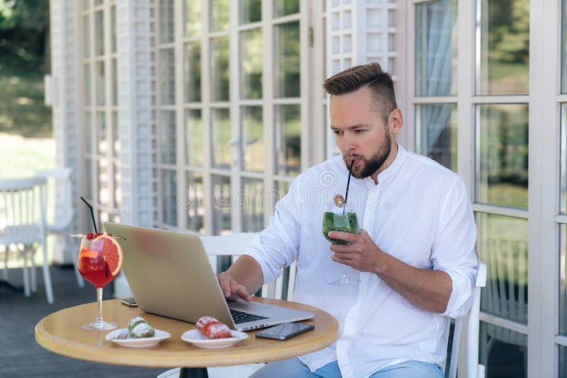 Un hombre de negocios está trabajando en su ordenador portátil, esperando a sus socios en una tabla en un café, bebiendo un cócte foto de archivo libre de regalías Un hombre de negocios está trabajando en su ordenador portátil, esperando a sus socios en una tabla en un café, bebiendo un cócte foto de archivo libre de regalías