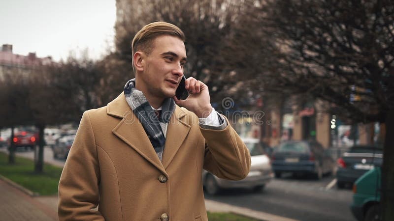 Un hombre de negocios joven en una capa que camina abajo de la calle en un tiempo nublado y que habla en el teléfono Cámara lenta