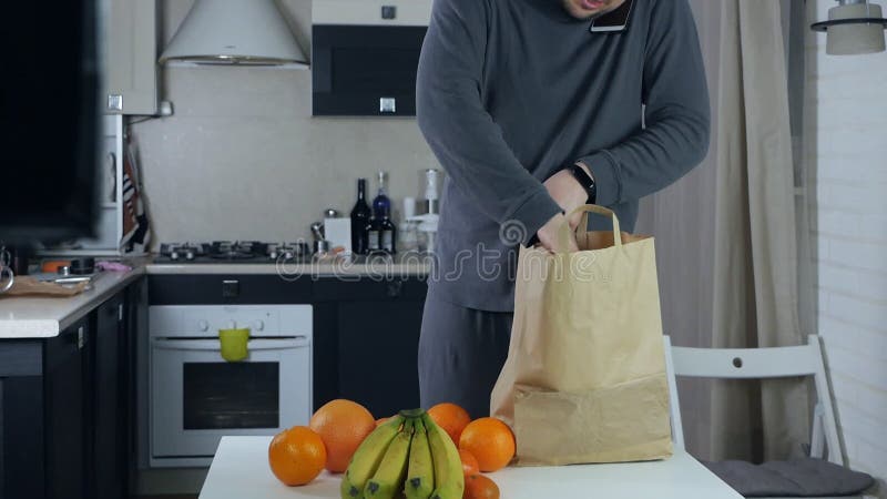 Un hombre desempaca alimentos frescos del mercado en la cocina y habla por teléfono metrajes