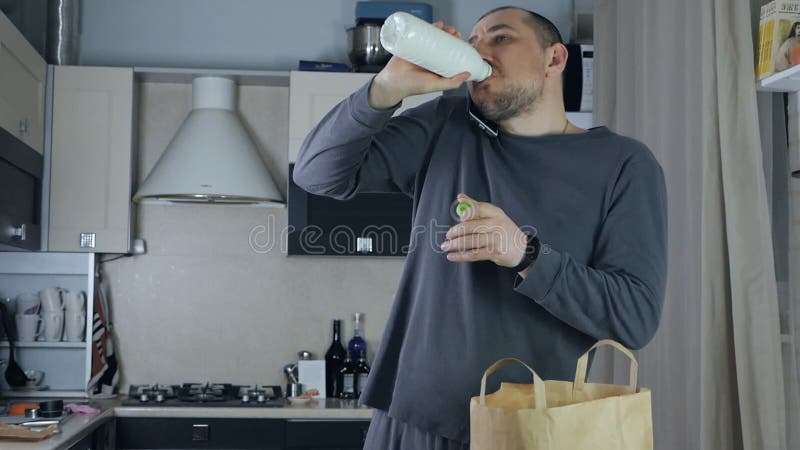 Un hombre desenvasa productos frescos del mercado en la cocina y habla por teléfono bebe leche fresca almacen de video