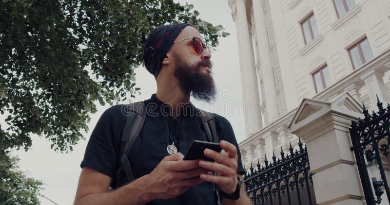 Un hombre elegante con barba navega por el paisaje urbano por su teléfono almacen de video