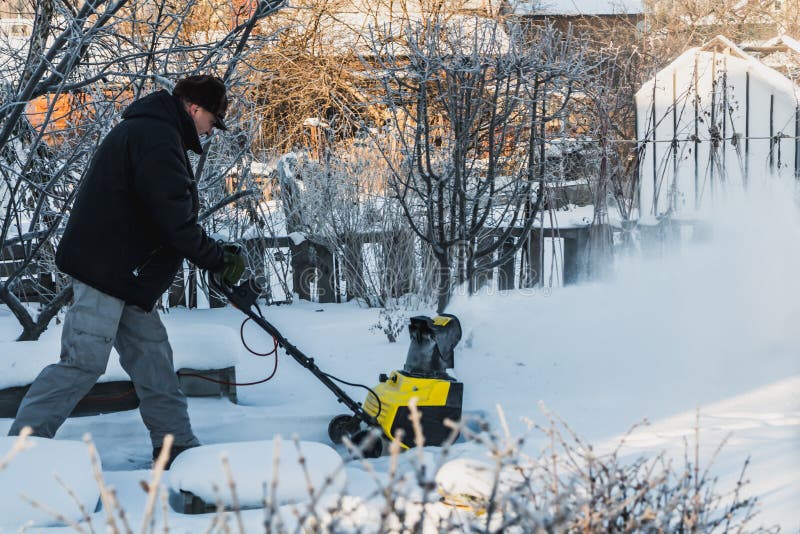 Un hombre en una chaqueta negra y los pantalones grises está cepillando la nieve blanca con el lanzador de nieve eléctrico amaril fotografía de archivo Un hombre en una chaqueta negra y los pantalones grises está cepillando la nieve blanca con el lanzador de nieve eléctrico amaril fotografía de archivo