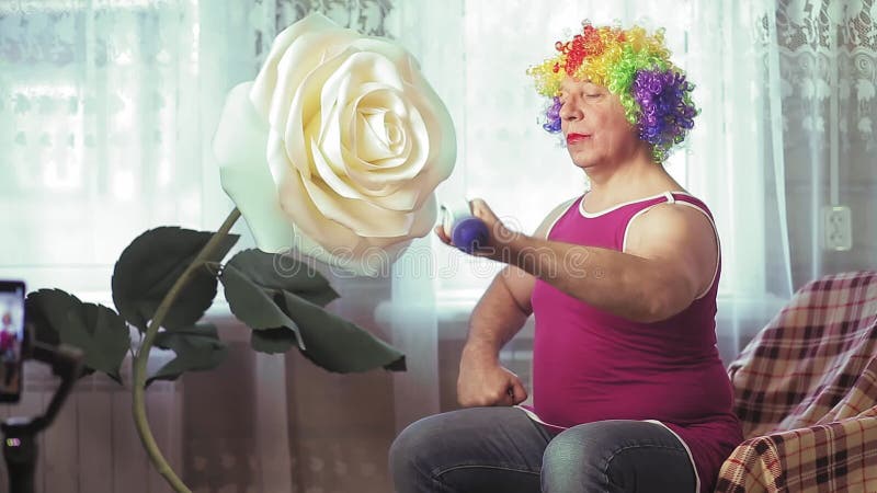 Un hombre gracioso en una peluca multicolor con maquillaje se sienta en un sillón y sacude a los bíceps de dumbbell.