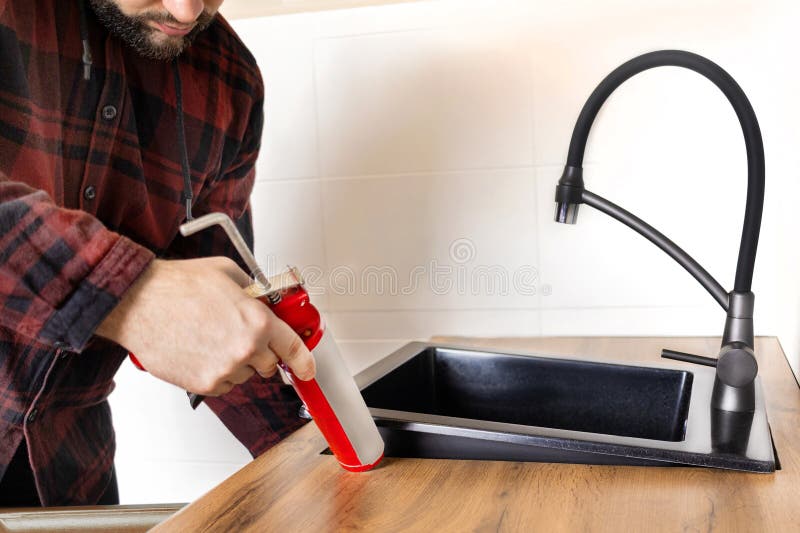 A man installs a black stone sink into a kitchen countertop. Worker seals up the kitchen sink with a sealant using a construction sealing gun close up. A man installs a black stone sink into a kitchen countertop. Worker seals up the kitchen sink with a sealant using a construction sealing gun close up