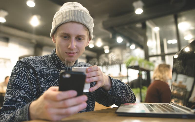 Un hombre joven positivo goza de un smartphone en un café mientras que trabaja para un ordenador portátil imagen de archivo Un hombre joven positivo goza de un smartphone en un café mientras que trabaja para un ordenador portátil imagen de archivo