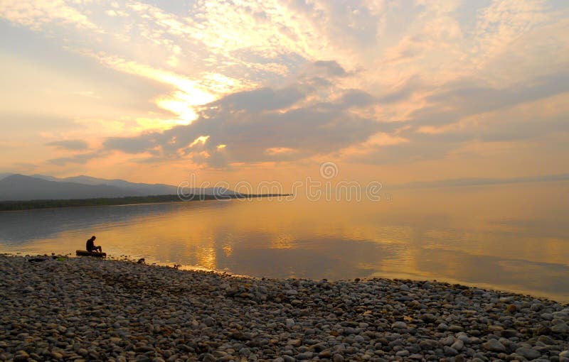 Un hombre que se sienta solamente en un registro en sobre el borde de un mar, silencio del amanecer en el lago