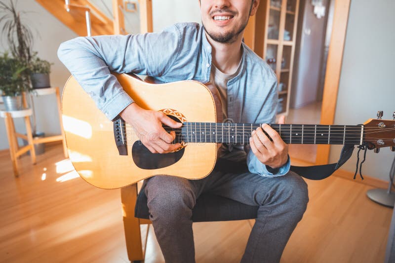 Un hombre solo en medio de una sala iluminada por el sol disfruta tocando una hermosa guitarra acústica de madera y componiendo nu imágenes de archivo libres de regalías