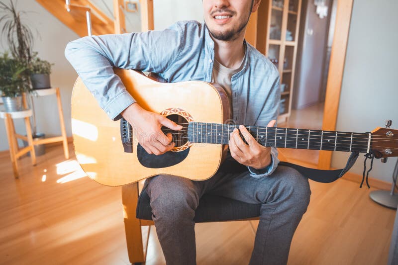 Un hombre solo en medio de una sala iluminada por el sol disfruta tocando una hermosa guitarra acústica de madera y componiendo nu imagenes de archivo