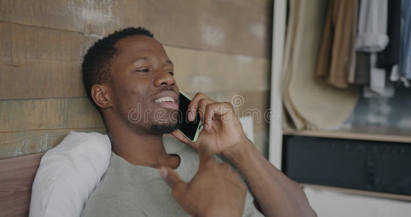 Un homme américain d'afrique heureux parlant sur un téléphone portable couché dans un appartement moderne banque de vidéos