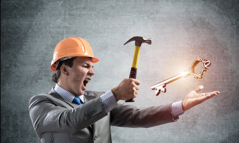 Furious businessman going to crash with hammer steel key. Young handsome contractor in business suit and safety helmet standing on grey wall background. Financial crisis in real estate. Password hack. Furious businessman going to crash with hammer steel key. Young handsome contractor in business suit and safety helmet standing on grey wall background. Financial crisis in real estate. Password hack