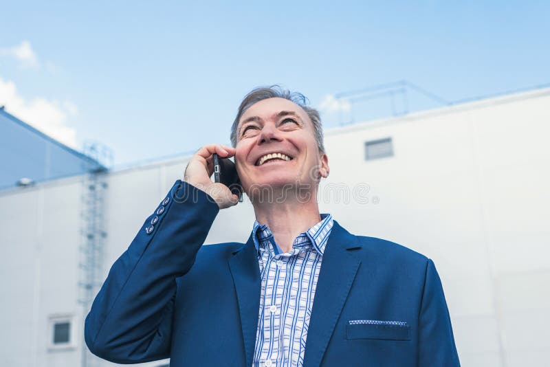 Un homme d'affaires d'âge moyen en costume bleu riant en appelant par téléphone de sa voiture sur le parking privé de la grande u photographie stock Un homme d'affaires d'âge moyen en costume bleu riant en appelant par téléphone de sa voiture sur le parking privé de la grande u photographie stock