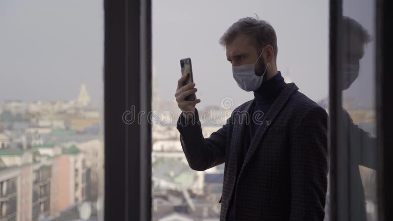 Un homme dans une veste et un masque parle via un lien vidéo sur le balcon d'un grand bâtiment du centre-ville